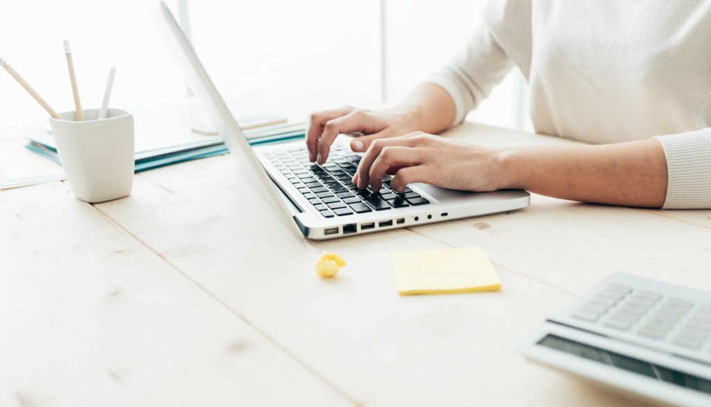 A woman in a white sweater is sitting at the desk and typing on her laptop.