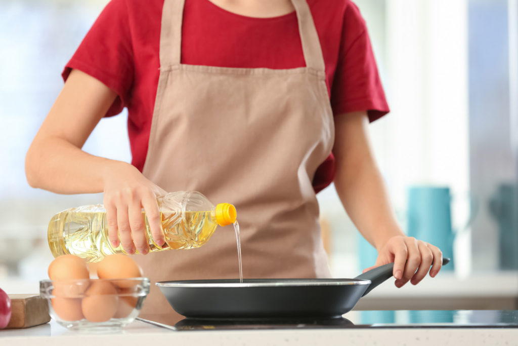 A woman is pouring edible oil from a plastic bottle to a pan on a stove.