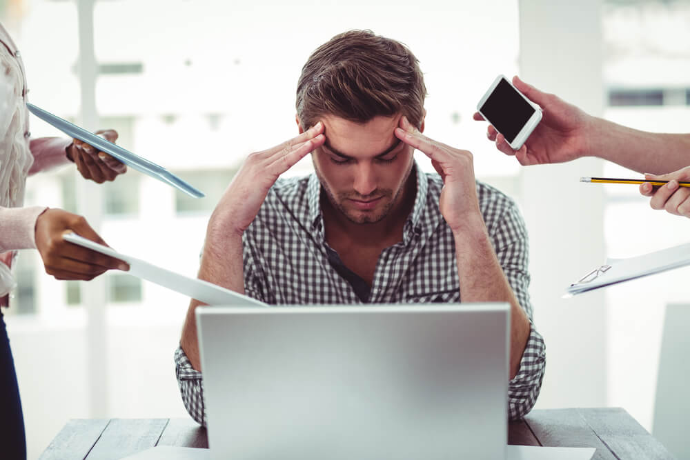 A man at work is sitting in front of a computer with his head in his hands because he’s under a lot of stress since they are pressuring him from all sides.