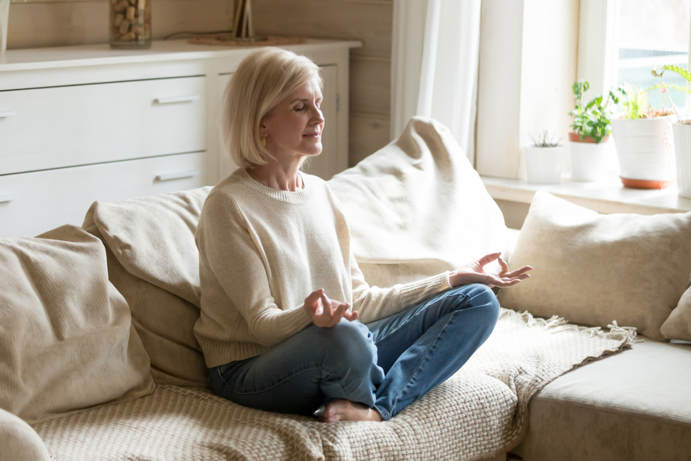 An elderly woman is meditating on a sofa in her home.