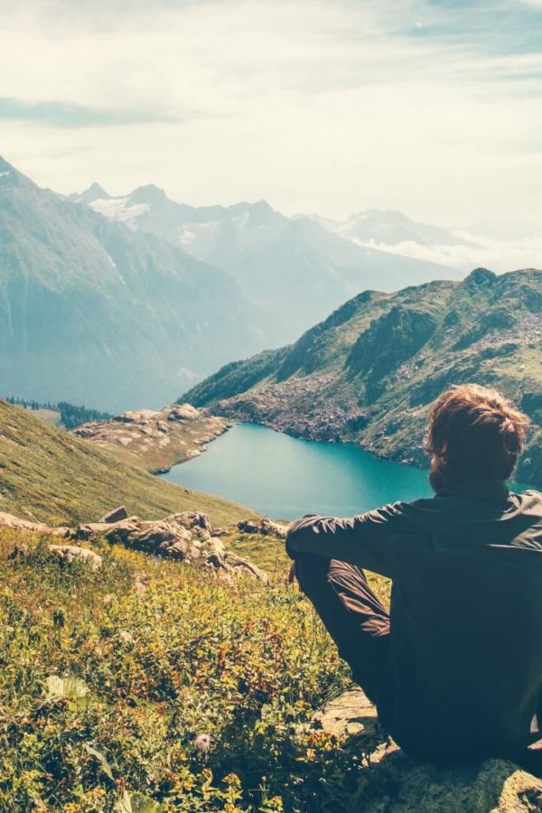 A traveler who is meditating by relaxing on a rock and admiring the magnificent view of the mountains and the lake.