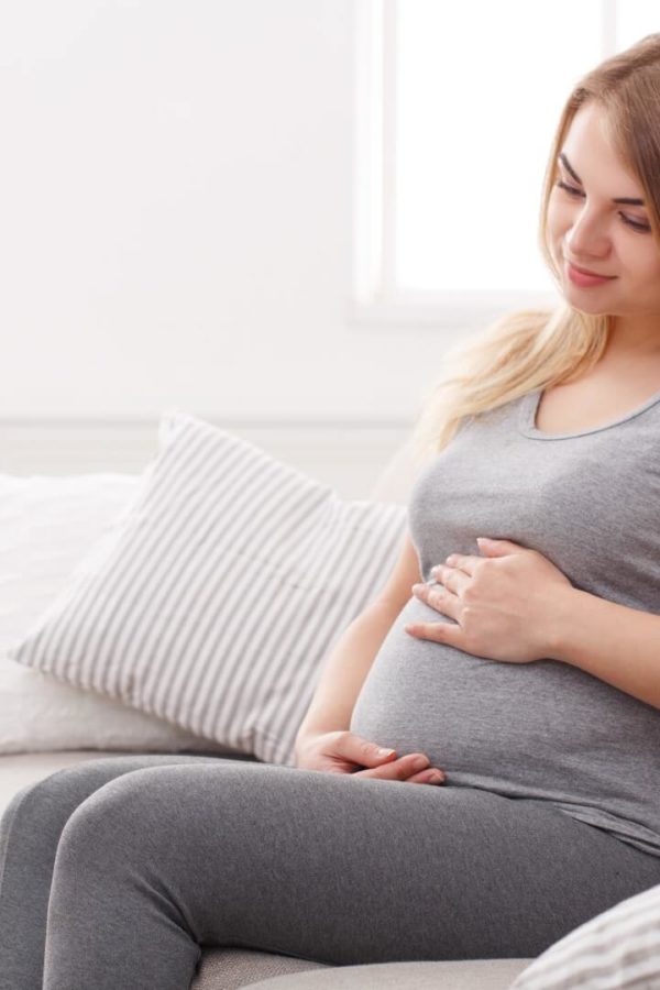 A woman wearing grey clothes is sitting at home on the sofa and caressing her stomach with both her hands.