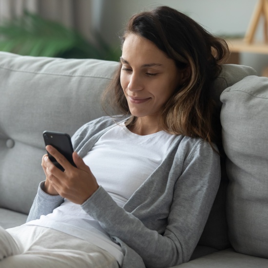 A smiling woman is resting on a comfortable sofa in her living room and browsing the Internet on a modern smartphone.