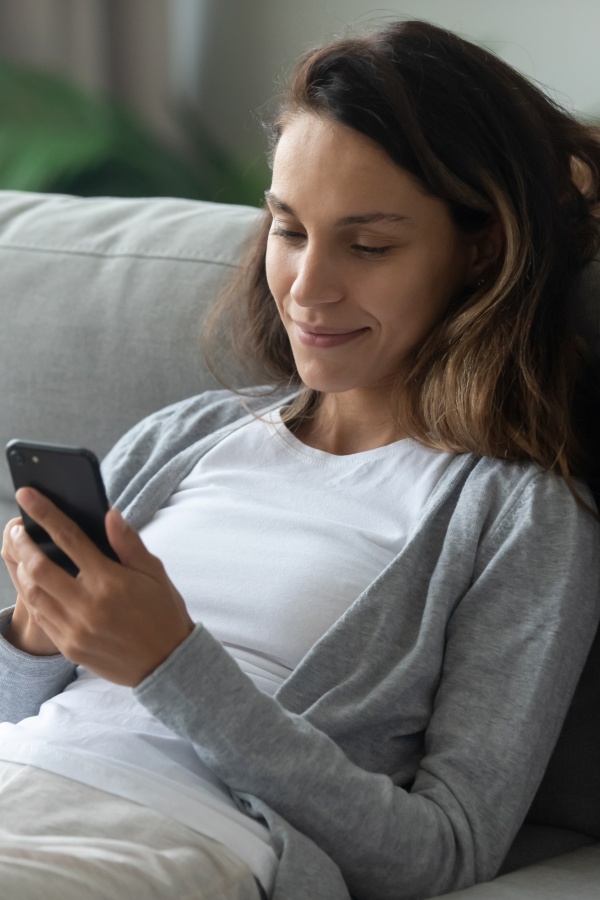 A smiling woman is resting on a comfortable sofa in her living room and browsing the Internet on a modern smartphone.