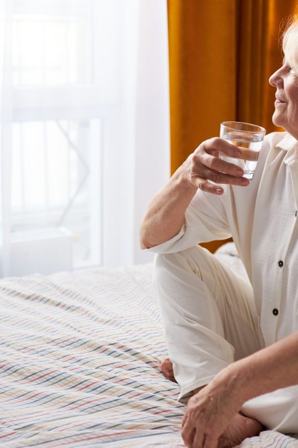 The older lady is sitting on the bed, drinking a glass of water and looking happily out the window.