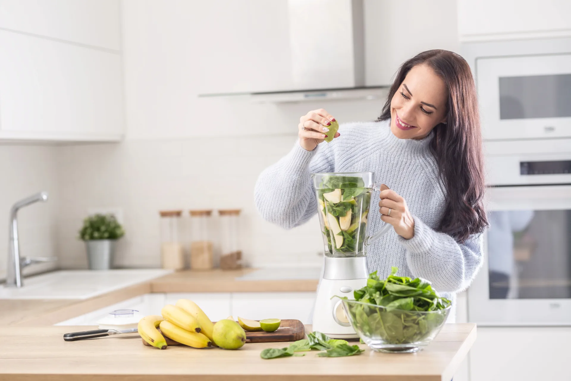 Ženska v svetlo modrem puloverju pripravlja zelen smoothie v kuhinji. Sestavine, kot so špinača in avokado, so v blenderju, medtem ko drži limeto, ki jo stiska. Na pultu so sveža zelenjava in sadje